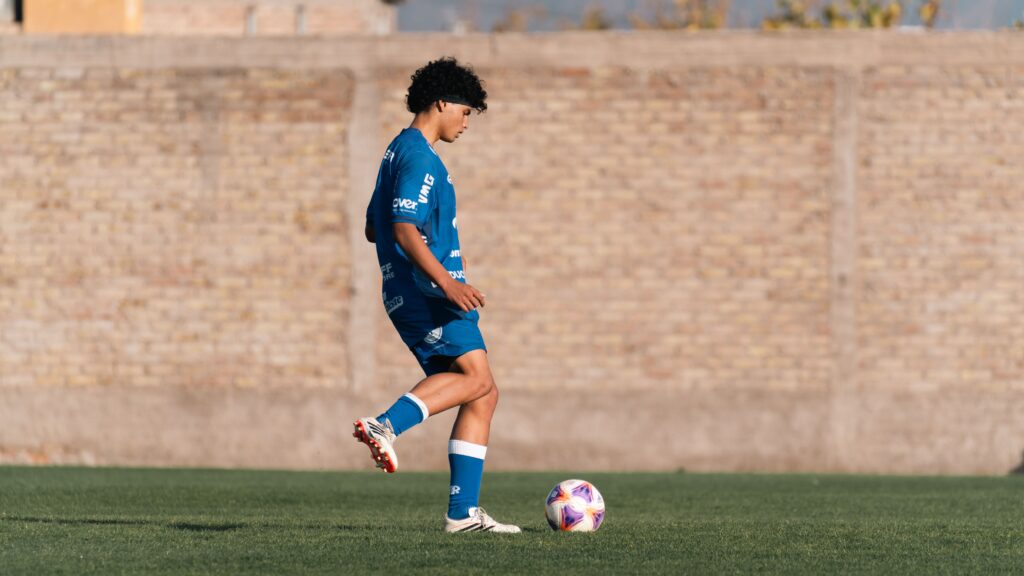 A teenage soccer player in blue kit practicing on a green field by a brick wall.